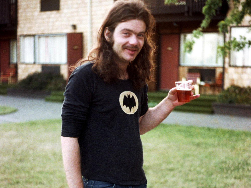 Roky Erickson in black t-shirt holding cup of beer in front of apartment building
