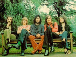 Early Fleetwood-Mac, with Peter Green in center, sitting on a bench with green grass and green trees