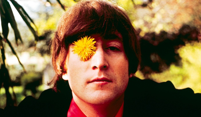Close-up of John Lennon wearing black jacket and red shirt with yellow flower on eye and blurred foliage in background