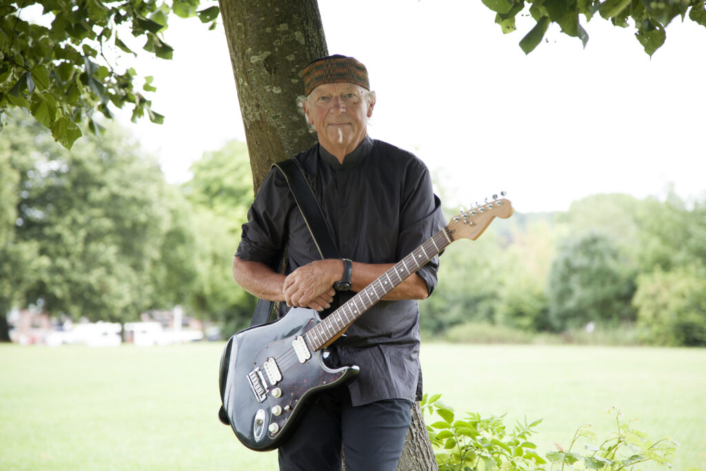 Martin Barre in black short-sleeved shirt and black cap leaning against tree with electric guitar hanging from his shoulders and field of grass and trees in background