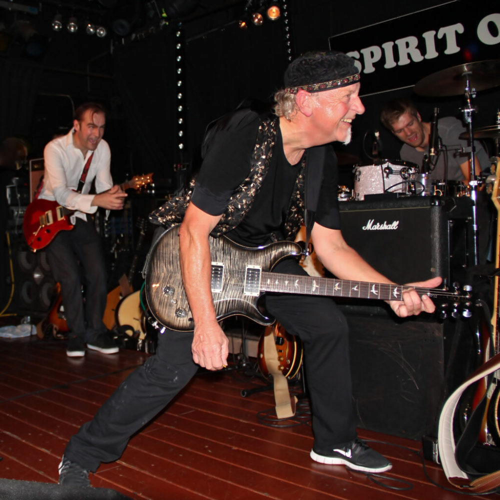 Photo of Martin Barre in black slacks, shirt, and cap strumming guitar while crouching and performing live onstage with musicians in background