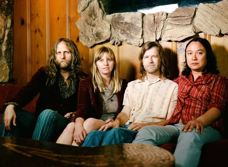 Sugar Candy Mountain's four members sitting closely on a couch looking at camera with wood wall behind them.