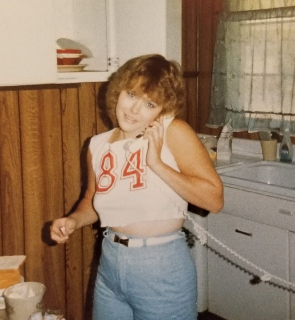 Young woman wearing a white half shirt with the number 84 on it and light blue shorts while on the phone (with a cord!) in what looks like an apartment
