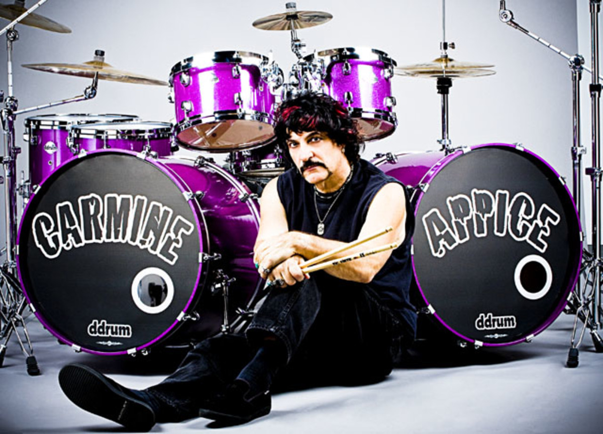 Carmine Appice sitting on floor with sticks in hand and purple highlights in his otherwise black hair in front of large purple drum kit with "Carmine" etched on one bass drum and "Appice" etched on the other.