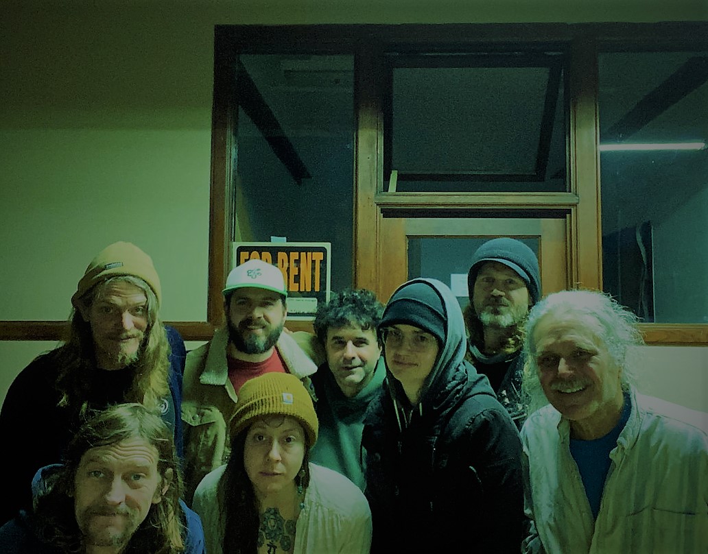Shining Realm band pic of eight members huddled in the hallway of an office building with a "For Rent" sign taped onto window behind them.