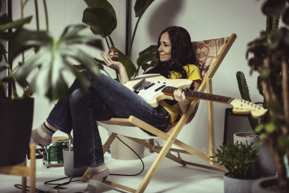 Juleah sitting in wooden recliner with white guitar in lap and house plants around her