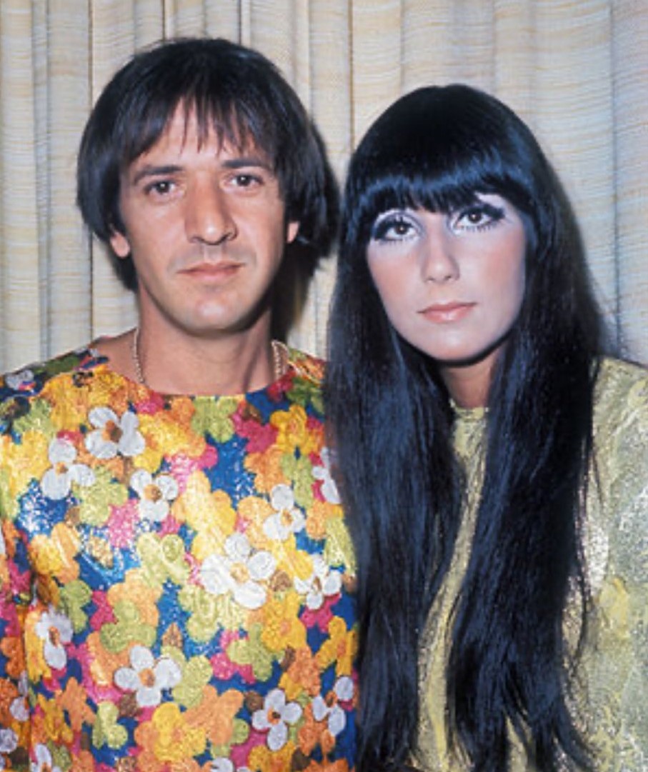 Sonny and Cher standing together with Sonny in multi-colored floral shirt