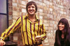 Sonny Bono in yellow and brown striped shirt with Cher outside by brick wall