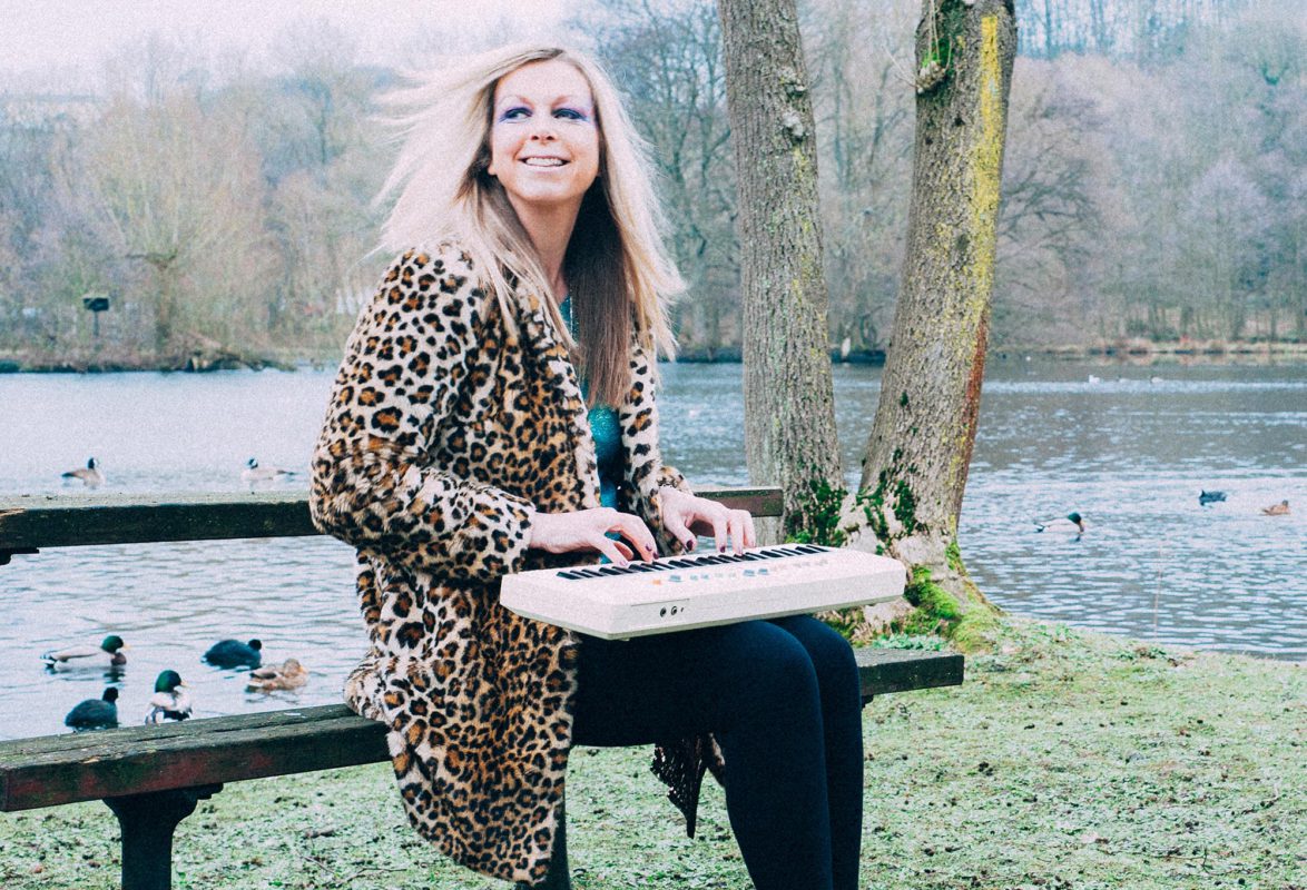 Jane Weaver wearing a leopard-skin jacket playing a keyboard on a park bench with green grass, trees, and a body of water behind her.