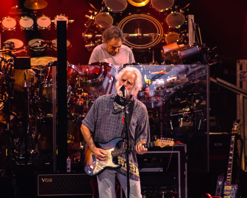 Bob Weir playing guitar and singing onstage with Mickey Hart drumming in background