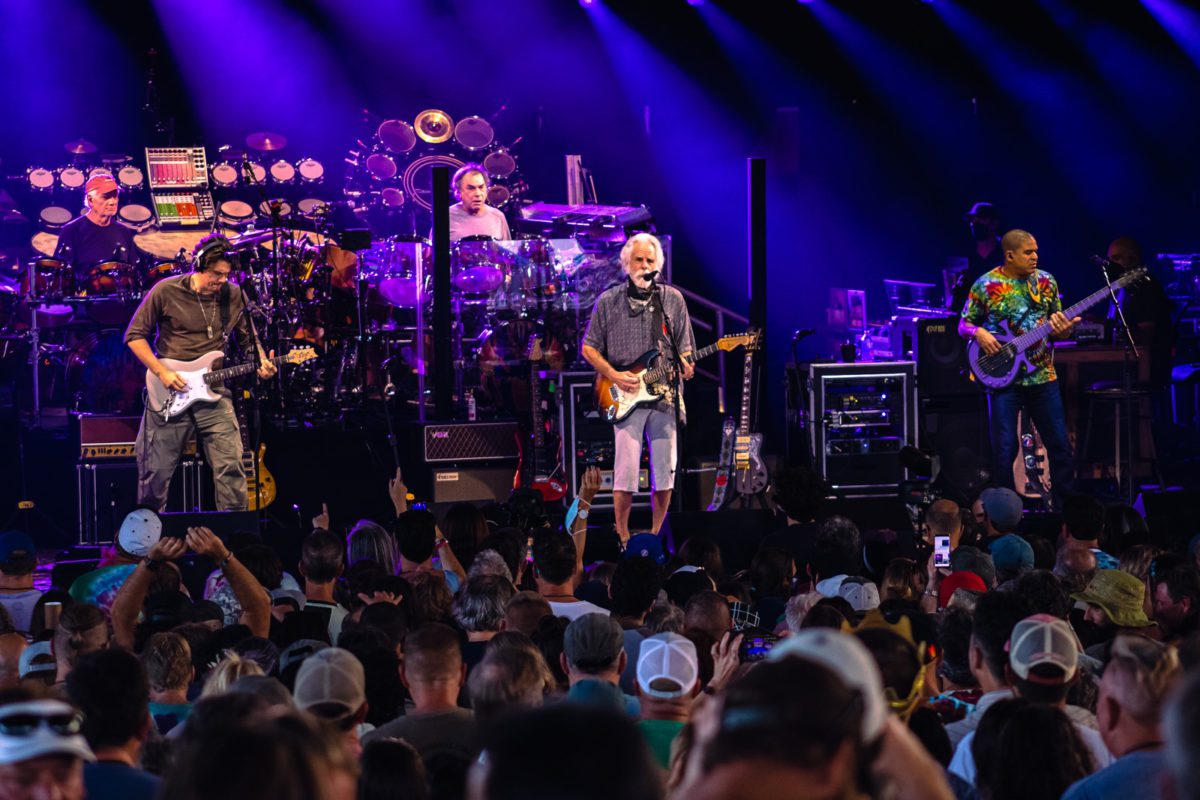 Dead & Company performing onstage in front of blue and purple background with the backs of many heads in the foreground