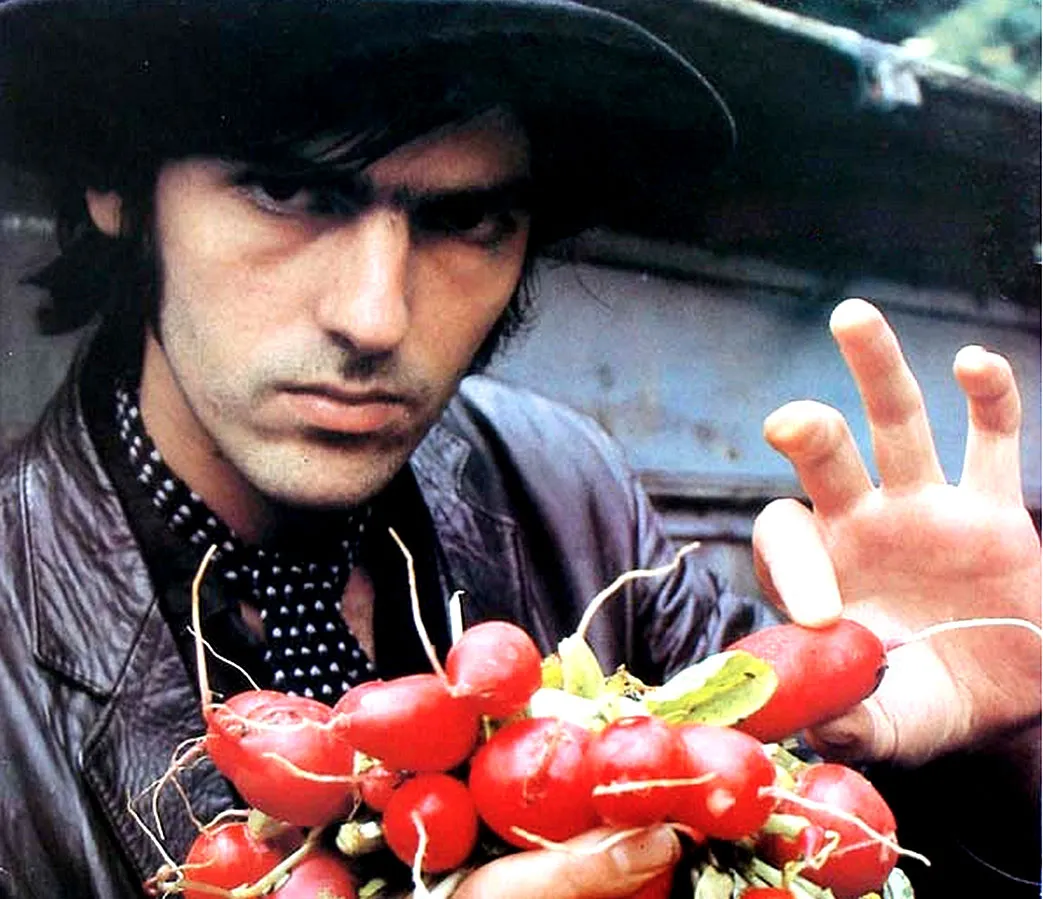 Robyn Hitchcock in leather jacket, polka dot tie, and hat holding red chili peppers