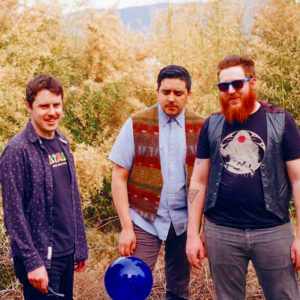 The three young men of Sun Voyager Band standing outside with forest trees in background