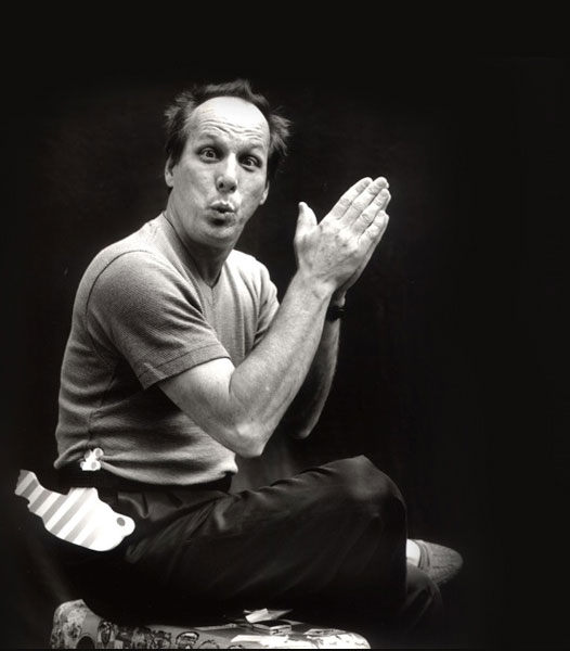 Adrian Belew sitting on floor with hands together and surprised look on face