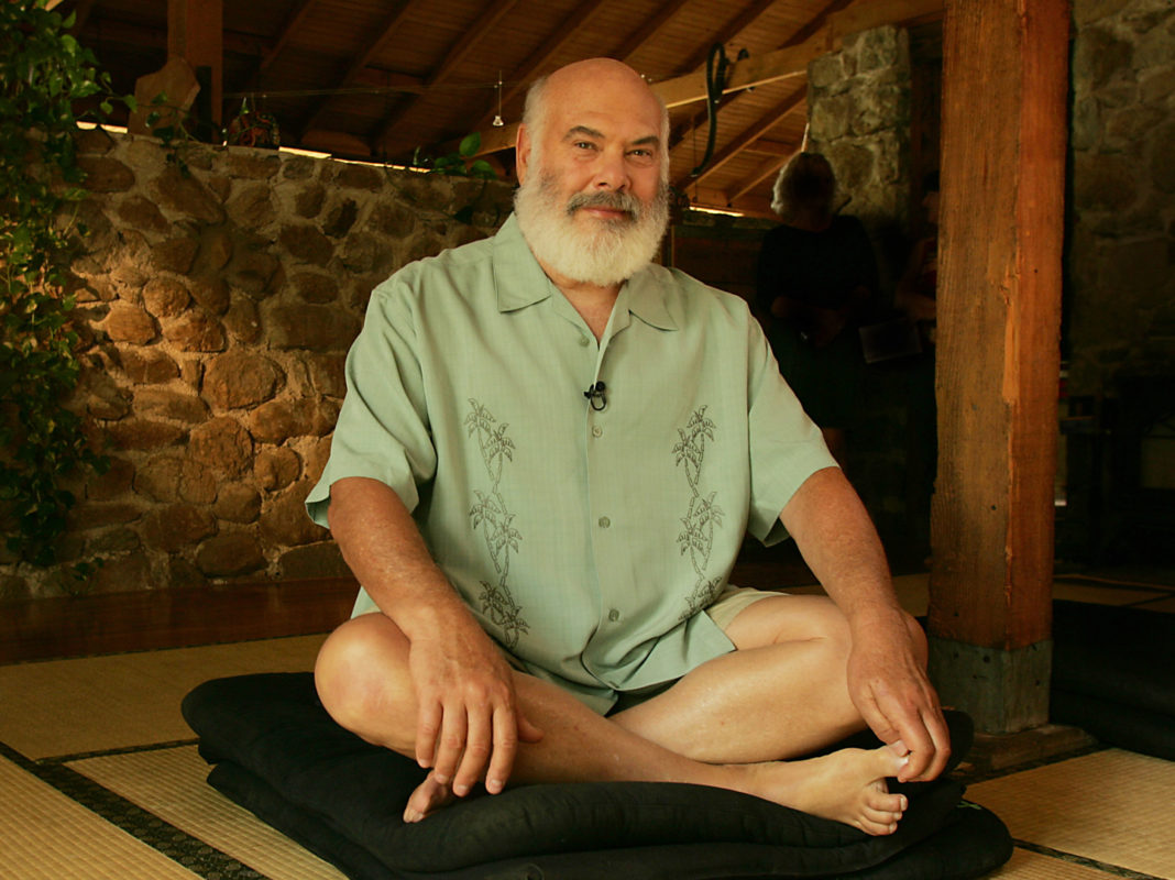 Dr. Andrew Weil casually sitting cross-legged on a black cushion in a hut