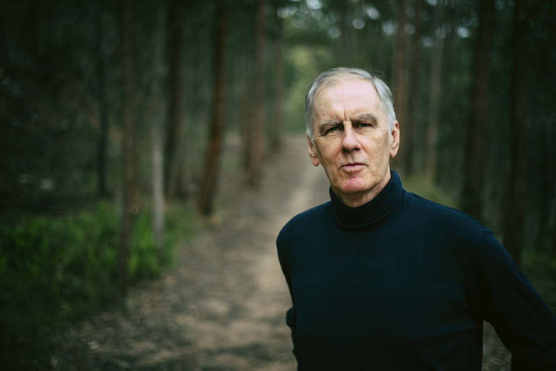 Robert Forster pictured in black long-sleeve shirt with a dark green forest in the background
