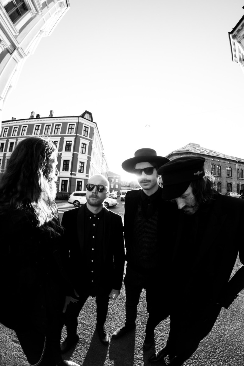 Black and white fish-eye photo of the four band members, all in black, standing on a street corner with old buildings in the background