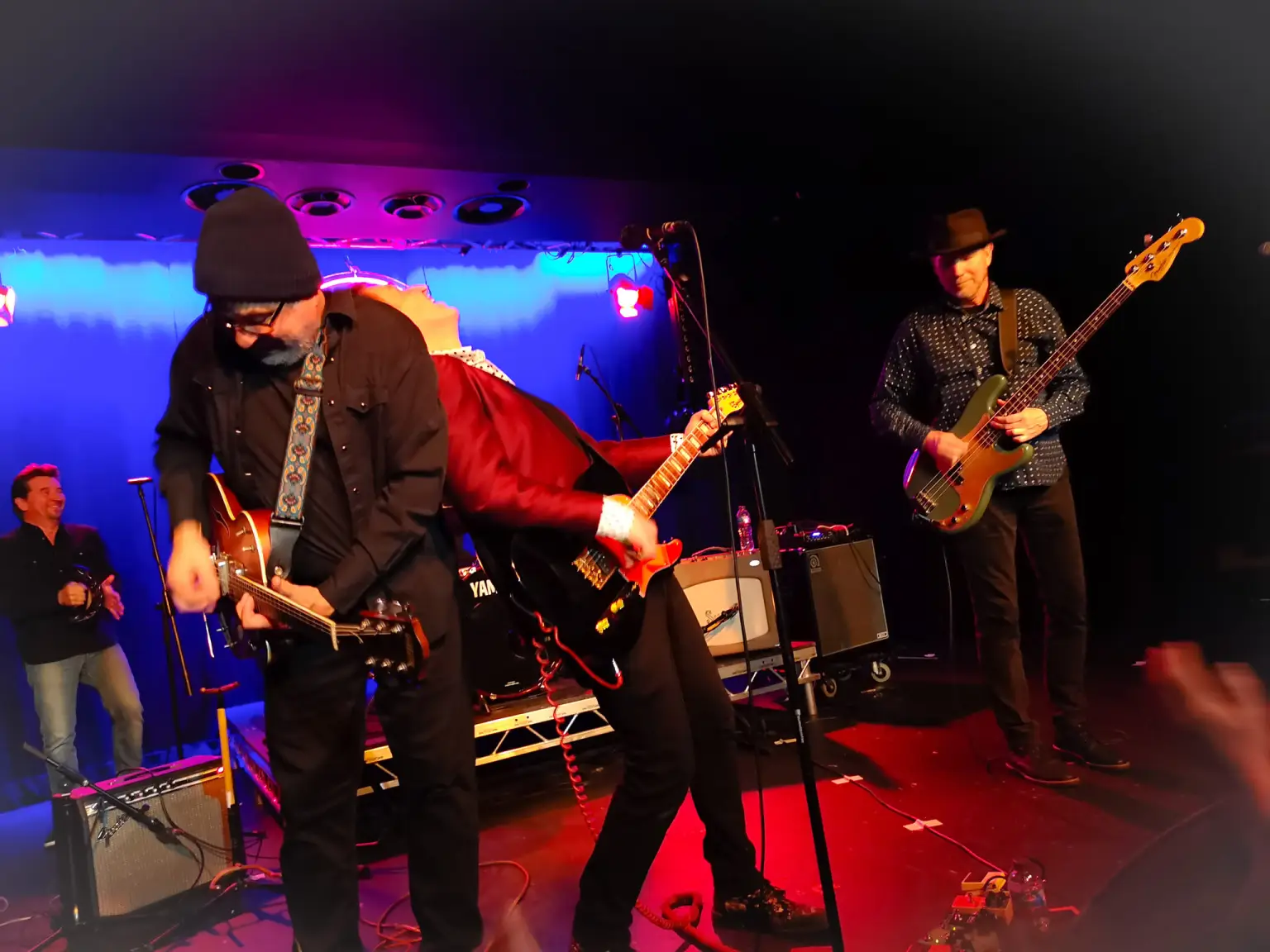 Guitarist in red coat and black slacks leaning up against guitarist Matt Piucci in all black with rest of band performing in background with red light on stage floor and blue and purple lighting behind them.