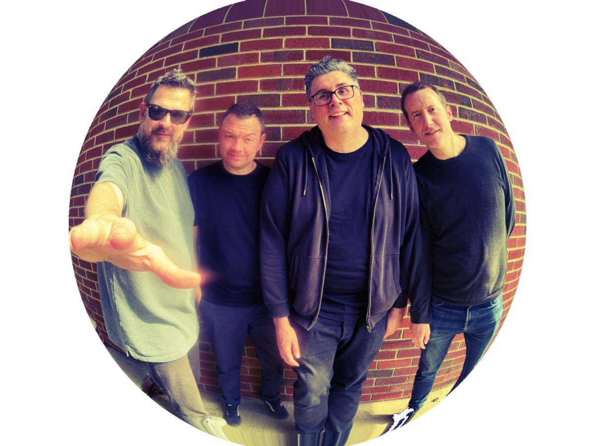 All four members of The Fierce and the Dead in casual attire standing in front of a red brick wall with a fish-eye lens effect.
