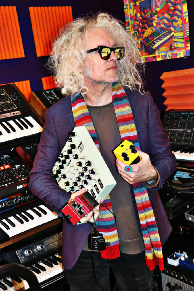 Anton Barbeau wearing cold weather clothes and a long, colorful scarf with keyboard in hand standing in front of larger keyboard