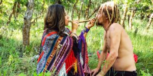 Woman with long brown hair in bright, multi-colored shawl holding pip up to nose of man with dreadlocks in a green field with trees in the background