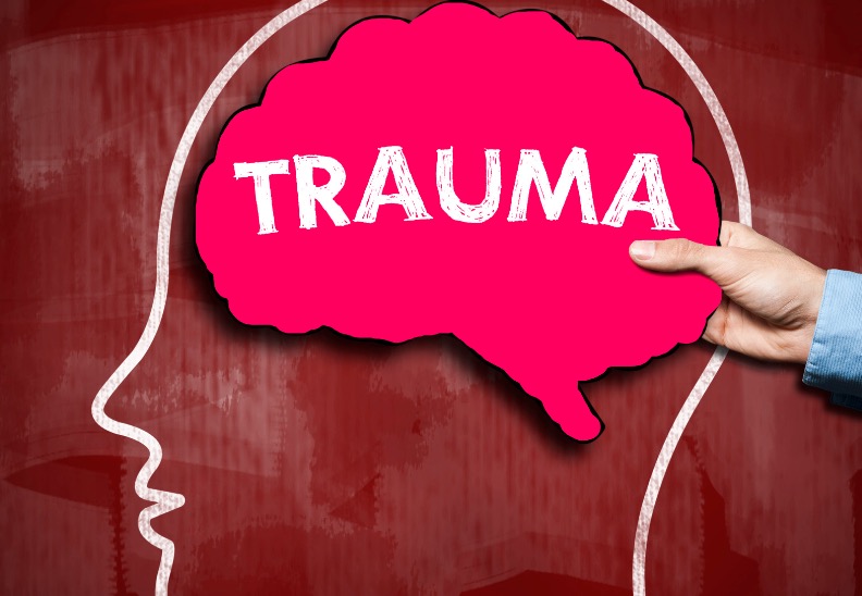 Photo of a chalk outline of a a person's profile with a hand holding a red brain-shaped card with the word "Trauma" over where the brain would be on the chalk person.
