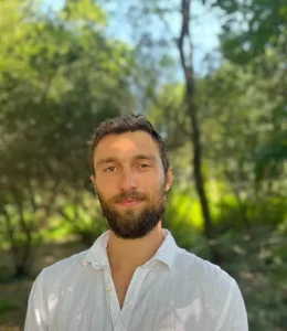 Young, bearded man in collared white shirt outside with green trees and grass behind him.