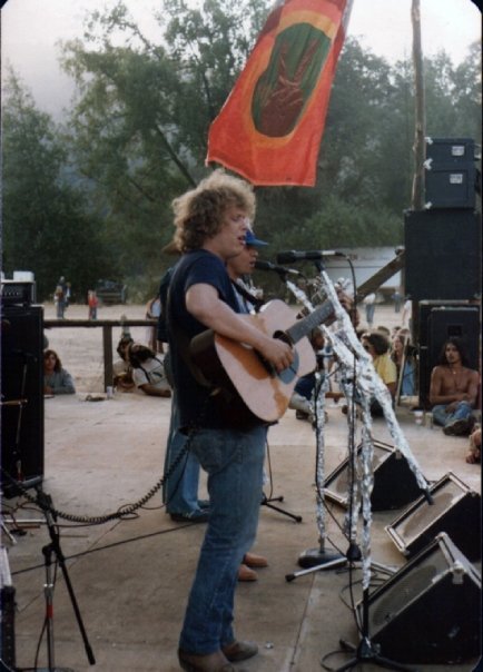 Barry Melton playing acoustic guitar outside with green trees in background
