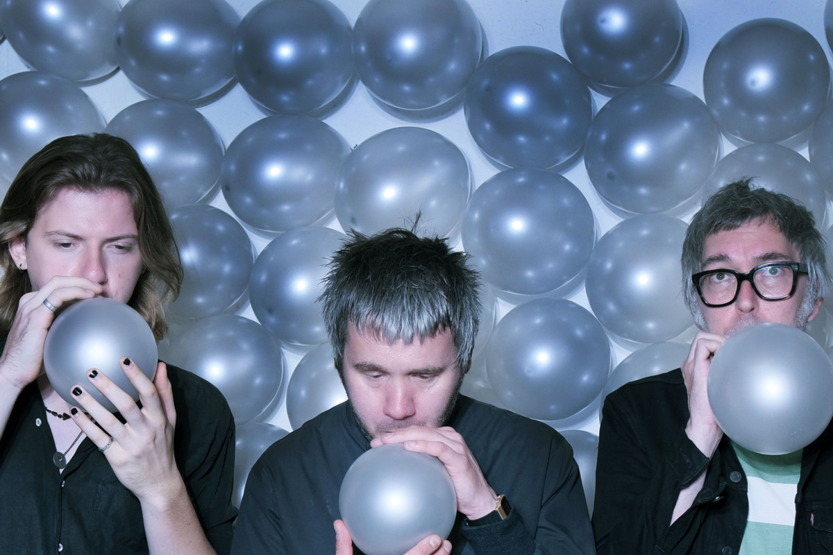 Three men in dark suits blowing up silver balloons with other silver balloons behind them on the wall