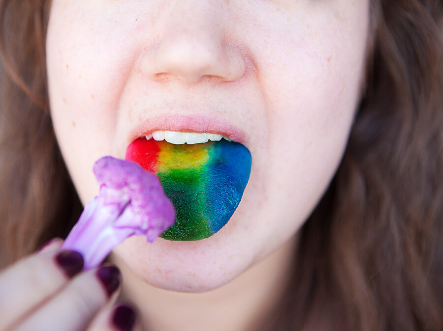 Close-up photo of a young woman putting purple cauliflower in her mouth while her tounge has multi-colored stripes