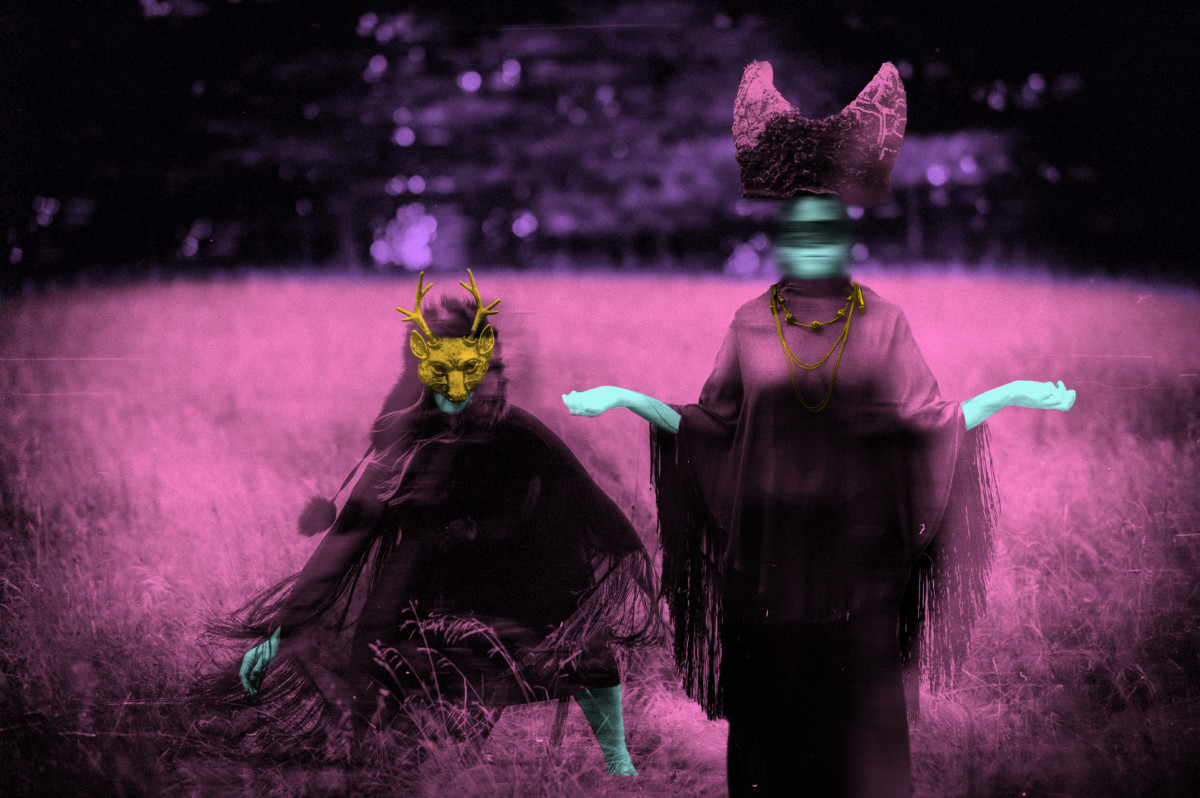 Blurred photograph of two individuals in a pink field, one wearing an animal mask and sitting with the other standing with arms outstretched-looks psychedelic.