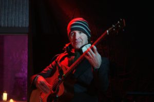 Malcolm Bruce wearing black suit and striped beanie playing bass under red spotlight with black background