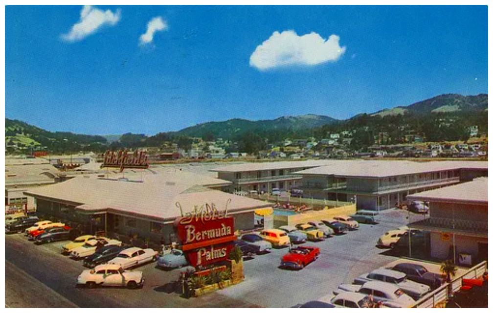 Aerial view of a moter hotel in the 1950s with old cars parked around it and a green mountain range behind it.