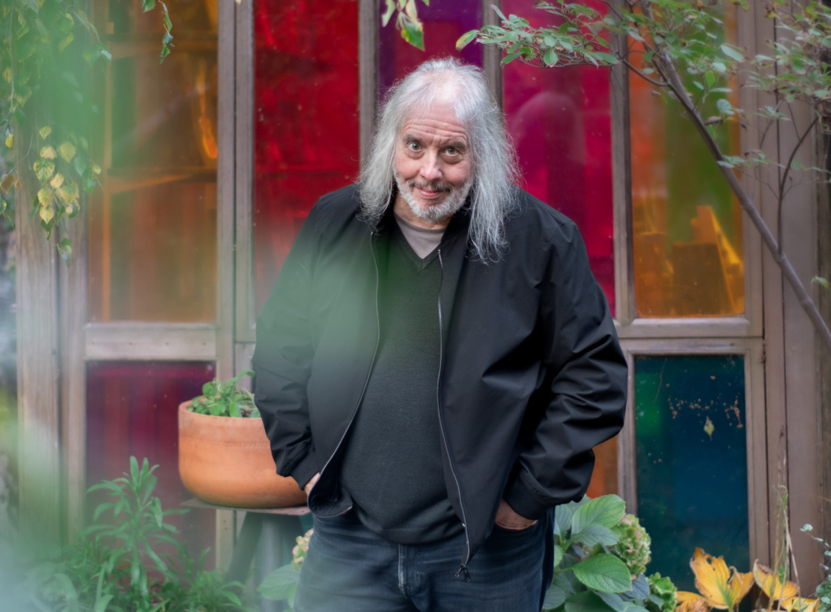 Man with long white hair wearing a black shirt and black jacket in front of mulit-colored windows