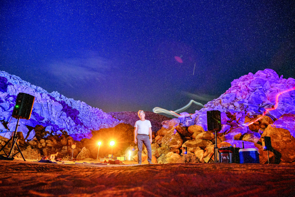 Man in jeans and white t-shirt standing outside in mountainous area with spotlights behind him.