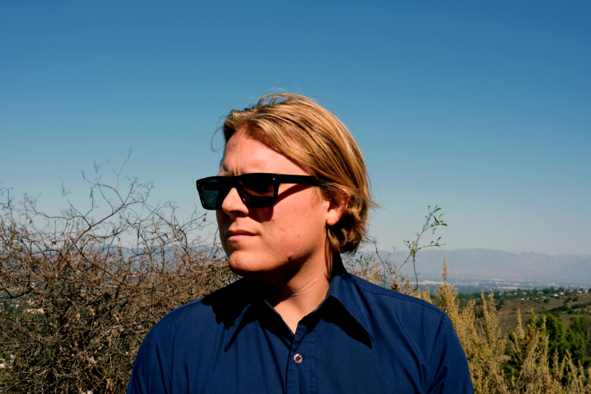 Photo of Ty Segall wearing a blue collared shirt and dark sunglasses outside in what looks like a field.