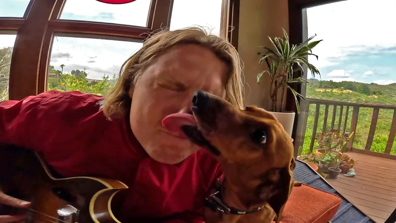 Ty Segall in his house with his dog, who is licking his face.