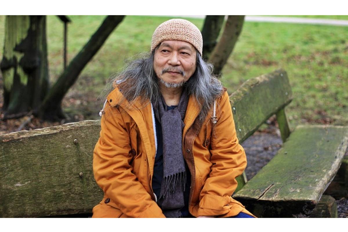 Photo of old Asian man with long graying hair in orange jacket sitting on park bench with green grass in background