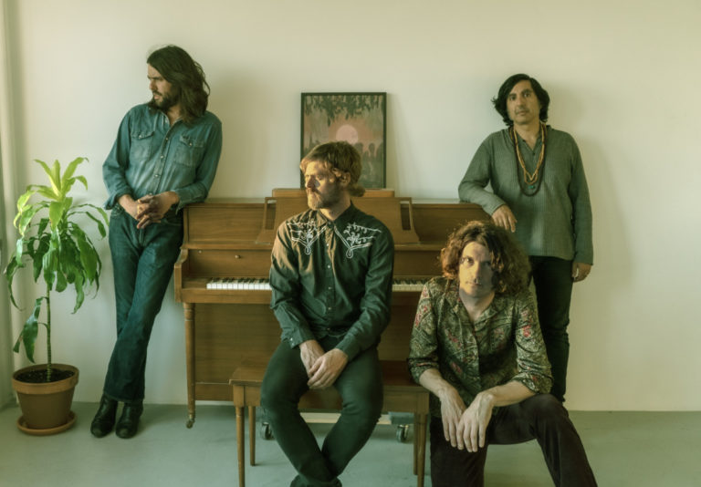 The four members of Elephant Stone posing for the camera with two members sitting and two standing against the wall by the piano.