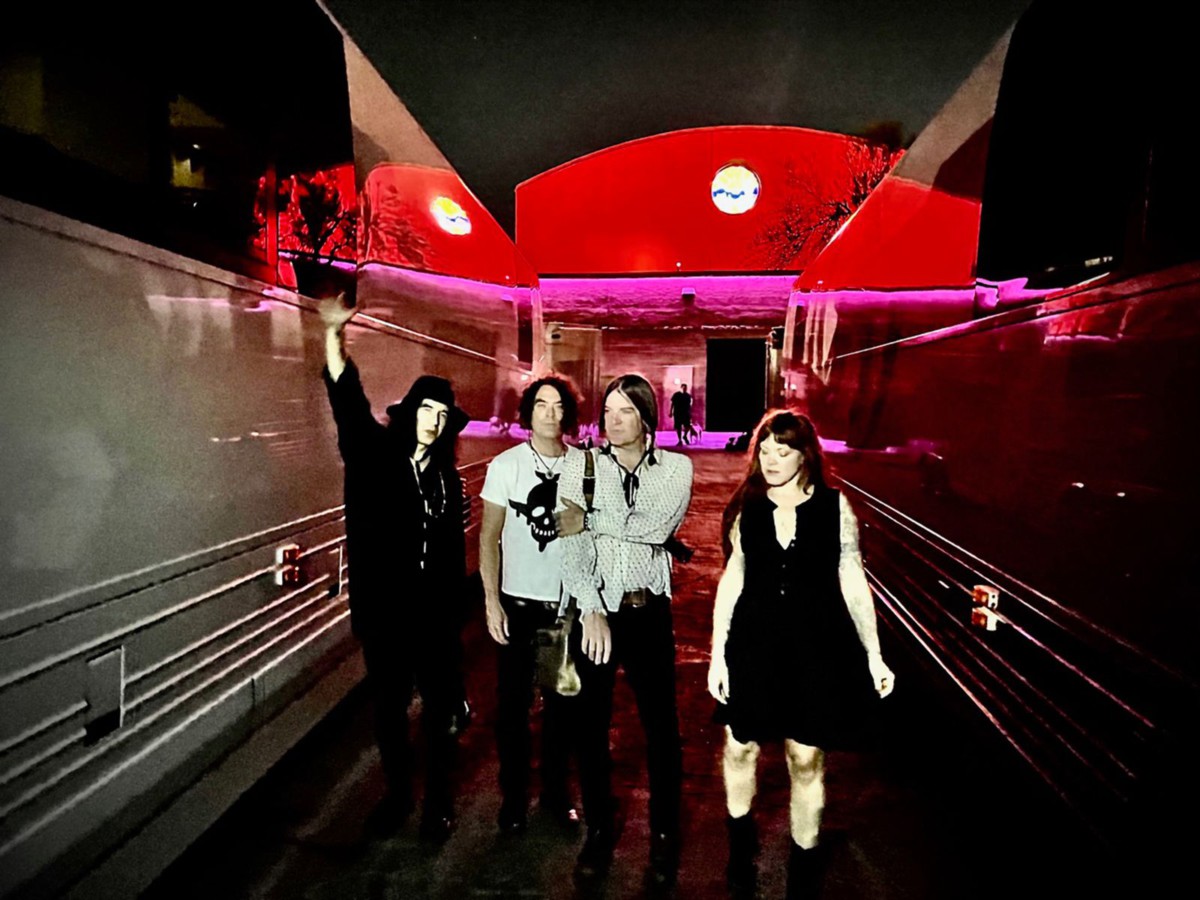 The four members of the Dandy Warhols walking outside on a wooded floor with red lights in the background