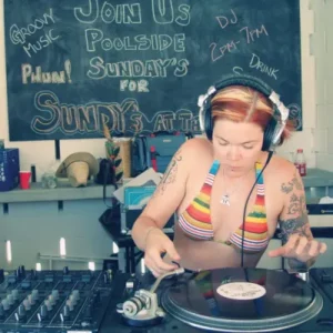 Woman in multi-colored bikini top wearing headphones looking down at vinyl record on turntable with mixer next to it