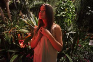 A young woman in a white gown stands holding her arms together and looking tenuous with a lush tropical background