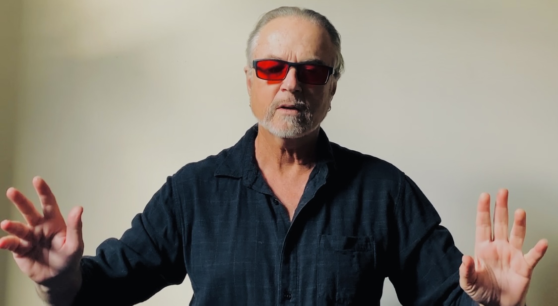 Photo of Steve Kilbey wearing red sunglasses with long-sleeved dark black shirt facing the camera with hands up at shoulder level.