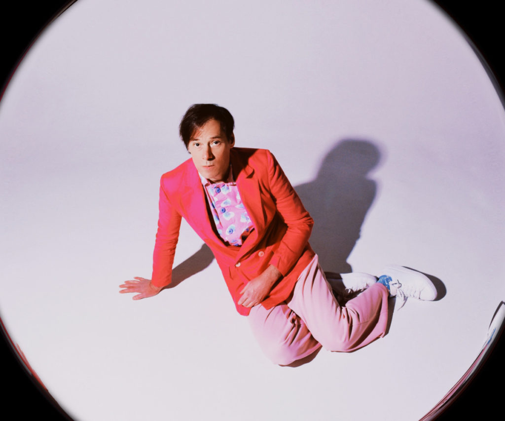 Overhead view of Kevin Barnes in in red blazer, pink slacks, and white shoes sitting on the floor looking up