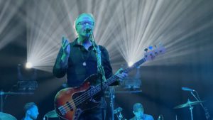Steve Kilbey with left hand on neck of bass guitar and right hand gesturing to crowd