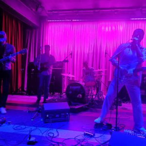The band Wand onstage with purple lighting on the four members with a pink background