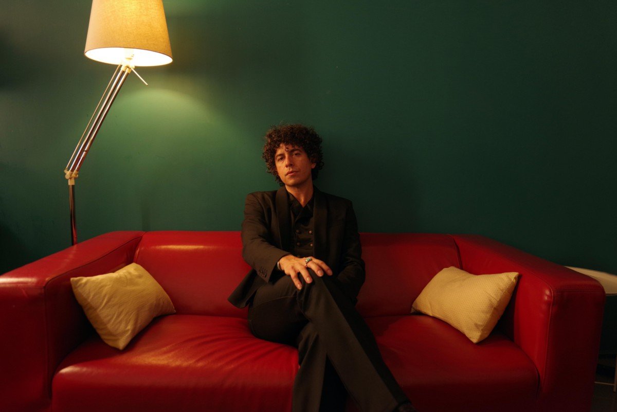 Brian Lopez dressed in black sitting on red couch in front of green wall with lampshade on