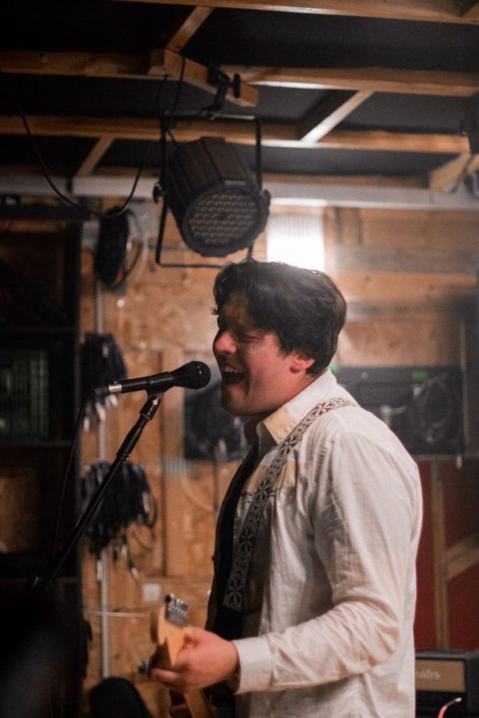 Photo of a man playing guitar and singing into a microphone in a studio with wood paneling