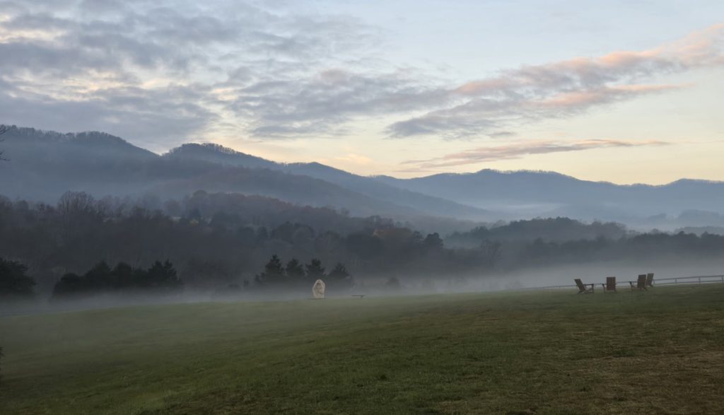 Photo of a foggy field with green grass and trees with mountains in the back and clouds above them.