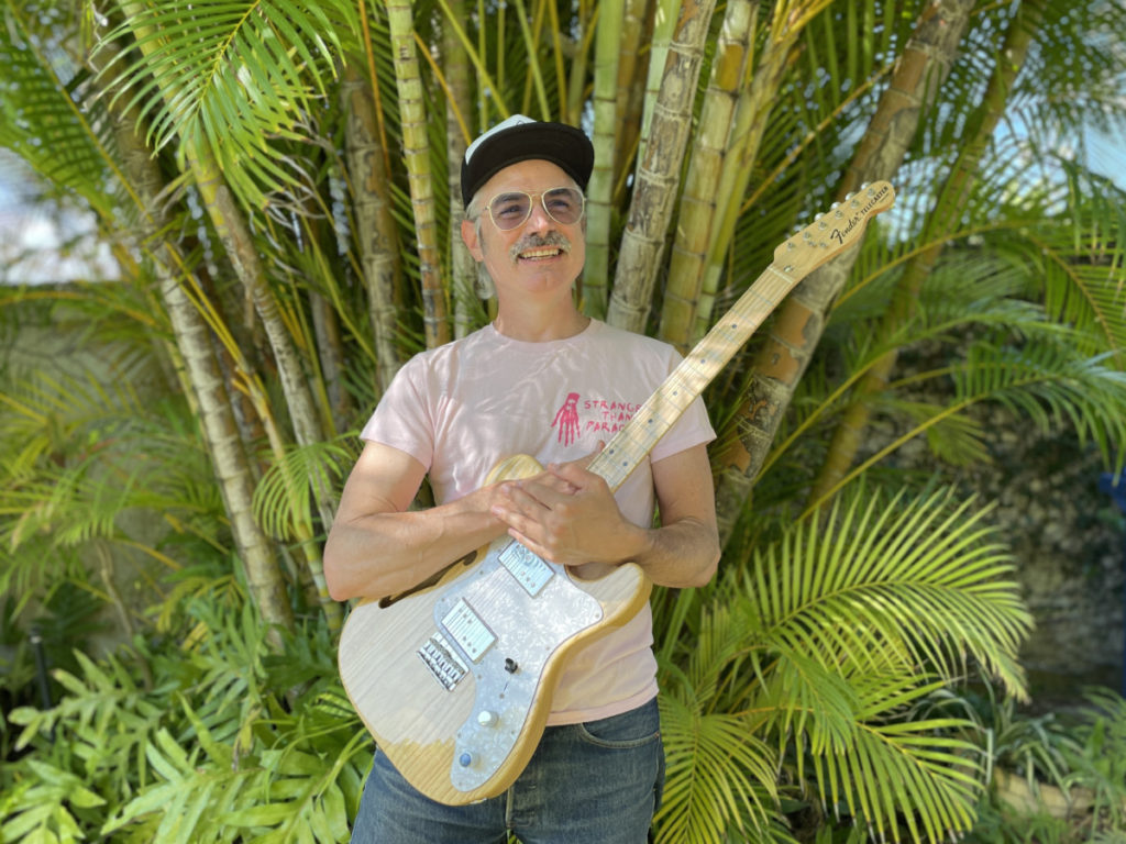 Photo of a middle aged white man holding an electric guitar in front of some greenery